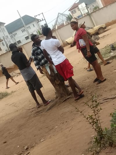 The scene is set in an unpaved dirt area within Umuokpu, Nigeria. In the foreground, the ground is light brown dirt with scattered small rocks and a low-growing green shrub on the right. In the midground, a dark brown tree stump is present.
Six individuals are visible. From left to right:
1. A person, possibly female, in a dark top and light headwrap, walks away from the camera towards the left.
2. A dark-skinned male, seen from behind, wears a black short-sleeved shirt, grey shorts, and light-colored flip-flops. He faces towards the right.
3. A dark-skinned male, facing right, wears a black and white plaid long-sleeved shirt and light-colored shorts. He holds a small, dark object in his left hand and stands to the left of the tree stump.
4. A dark-skinned male, seen partially from behind, wears a white short-sleeved t-shirt, red shorts with a white logo, and black flip-flops. He leans against the tree stump, facing towards the right.
5. A dark-skinned female, facing left, wears a red short-sleeved top, dark patterned shorts or skirt, and a patterned headwrap. She holds a white notebook or binder in her left arm and a pen in her right hand. She wears dark flip-flops.
6. A partially obscured dark-skinned male, wearing a light-colored cap and dark shirt, stands behind Person 5, near a yellow, dusty sedan car that is parked on the dirt path. He wears dark flip-flops.
In the background, a white multi-story building with a dark grey roof and multiple windows is visible on the left. A concrete wall topped with barbed wire runs across the mid-background. On this wall, a sign reads "BENSON EDUCATION CENTRE" and "JAMB." Overhead power lines and poles are also visible.
