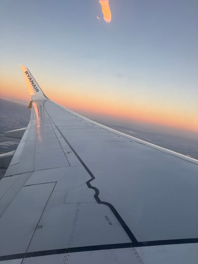 An aerial view shows the wing of a Ryanair aircraft during sunset. The wing extends from the lower left to the upper right of the frame. The Ryanair logo is visible on the wingtip. The sky transitions from a clear blue at the top to shades of orange and pink near the horizon. Below the wing, a hazy landscape suggests a city or town, likely Hart, United Kingdom, though no specific landmarks are identifiable. There are no people or specific actions depicted. The scene is static, focusing on the aircraft wing and the atmospheric conditions.