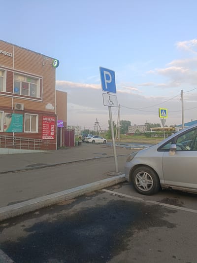 A silver car is parked in a marked parking space on a paved lot adjacent to a building with brown siding. A blue and white parking sign for disabled individuals is visible to the left of the car. The building has several windows and signage, including a sign that reads "МЕБЕЛЬ" (Furniture) and another with the "OZON" logo. A pedestrian crossing sign is located further down the street. The sky is partly cloudy and the scene appears to be in a city. This is the parking lot of the "Planetа" shopping center located at 89, 50 Let Oktyabrya Street, Bratsk, Russia.