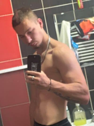 An overhead-oriented mirror selfie captures a shirtless young adult male in a bathroom in Vetren Dol, Bulgaria. The man, with short dark hair, is seen from the chest up, facing slightly away from the camera's perspective, looking towards his reflection. He holds a black smartphone in his right hand to take the photo, with a silver ring visible on his right ring finger. A silver chain is worn around his neck, and tattoos are visible on his left forearm.
The bathroom features dark grey/black tiled walls with white grout lines in the upper section and bright red tiles with white grout lines in the lower section, separated by a white border. In the mirror's reflection, behind the man, a white towel hangs over a rack. Various bathroom items are visible, including a bottle with a yellow cap and a small red and green object, possibly a product tube, on a shelf. No discernible text is visible on any of the products or items in the reflection due to blurriness. The time of day or specific weather conditions are not evident from the indoor setting.