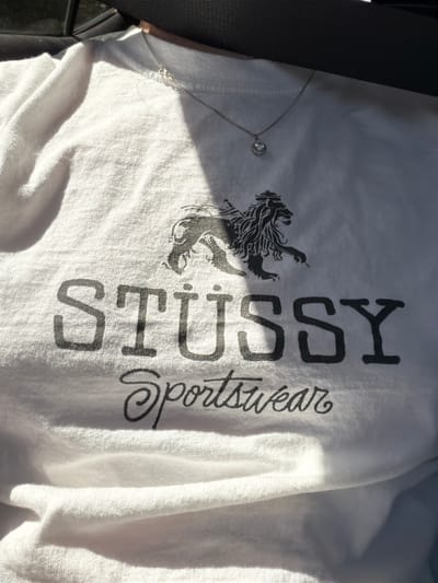 The image shows a white t-shirt with black graphics. The graphics include a stylized lion and the text "STÜSSY Sportswear". The t-shirt is draped over a dark surface, possibly the interior of a car. A silver chain with a heart-shaped pendant is visible on the t-shirt. The scene is lit by sunlight, casting shadows on the fabric. The location in Seaside, United States is not discernible from the image.