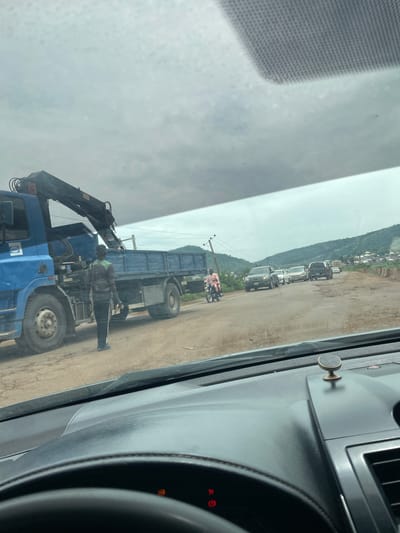 The image shows a street scene in Koton Karifi, Nigeria. The perspective is from inside a vehicle, looking out through the windshield. The day appears overcast, with a dull, diffused light suggesting it might be late morning or early afternoon, or perhaps a cloudy day.
The main focus of the image is a large blue truck with a crane attached. Two people are visible near the truck. One person, wearing a light-colored shirt and dark pants, is standing on the ground near the rear of the truck, appearing to be directing or observing. Another person, wearing a green reflective vest and dark clothing, is positioned on the truck itself, near the crane. This suggests some kind of operation is taking place involving the truck and its equipment, possibly loading or unloading.
In the background, a line of vehicles is parked along a dirt road. These appear to be mostly SUVs and cars. Beyond the road, a hillside is visible with scattered buildings and vegetation, indicating a semi-urban or rural setting. A motorcycle with a rider is also present on the road, adding to the sense of everyday activity.
The interior of the vehicle from which the photo is taken is partially visible, including a dashboard with some controls and a circular attachment on the dashboard. The overall scene depicts a moment of work or traffic on a road in Nigeria. There is no discernible text in the image.