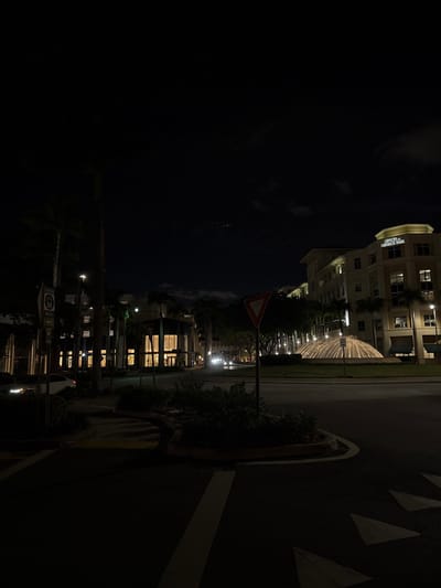 This night-time image captures a quiet street scene in Coral Gables, United States, characterized by urban architecture and illuminated features. The main subjects are multi-story buildings, a roadway, and landscaped greenery under a dark sky.
A prominent building on the right features modern architecture with numerous lit windows. Text clearly visible on this building identifies it as "OFFICES AT MERIDIAN PARK." Below this building, a distinctive, brightly lit water feature, possibly an architectural waterfall or light installation, creates a glowing arc that stands out against the darkness.
In the foreground, a dark asphalt road is marked with white triangular arrows, typically indicating a yield or directional instruction, along with solid white lines. A red triangular yield sign with a white border is also visible on a pole near the road. Streetlights dot the scene, casting light on the pavement and surrounding foliage.
The sky, occupying the left side of the frame, is largely dark, suggesting a clear night. A few faint specks of light are visible in the upper left, possibly distant stars or artificial lights. No people are present, and the scene is static, conveying a sense of calm and stillness typical of a city at night.