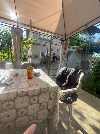 The image shows a sunny outdoor patio with a table covered by a floral tablecloth. On the table, there are cigarettes, a glass of amber liquid, and a container. A man is sitting on the stairs of a house in the background. A chair with a jacket draped over it is also visible. The environment is green with trees and some potted plants. The overall scene appears to be a casual outdoor gathering in Rogozen, Bulgaria.