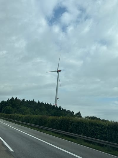 In Elz, Germany, a tall white wind turbine stands against a cloudy sky, its blades angled upward. The turbine is positioned on a forested hill, with dark green trees forming a dense line across the landscape. In the foreground, a multi-lane asphalt road runs diagonally from the lower left to the upper right corner of the image, bordered by a metal guardrail and a hedge. White lane markings are visible on the road. The overall scene depicts a rural or semi-rural environment with a focus on renewable energy infrastructure.