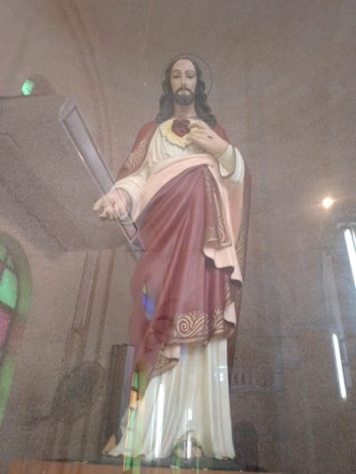 The image shows a statue of Jesus Christ in what appears to be a church or religious setting. The statue is standing, draped in white and maroon robes with golden decorative edges. A heart with a yellow halo is visible on Jesus' chest. The statue has long brown hair and a beard. The statue is housed in a display case with a stained-glass window and what looks to be modern rectangular lights on the sides. It is placed on a wooden platform. A blurred fan is located behind the glass. The surrounding location is likely the city of Villa Hayes in Paraguay.
