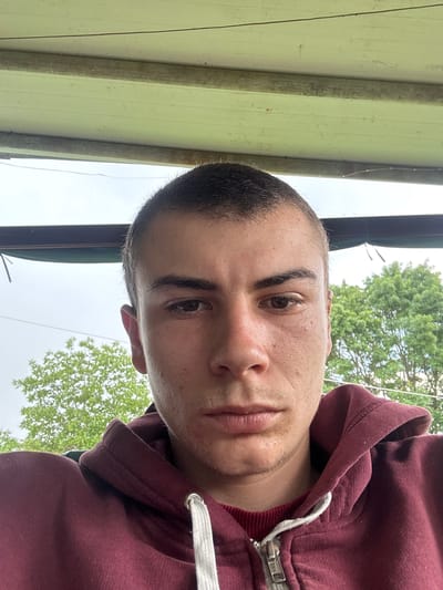 A close-up photograph depicts a young man wearing a maroon hooded sweatshirt with a white drawstring. His short hair is dark brown, and his eyes are brown. The background includes a green metal structure and trees, suggesting an outdoor location. The photograph does not definitively confirm the city of Dabovets, Bulgaria. The lighting is overcast.