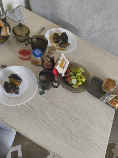 The image depicts a top-down view of a wooden table set with a meal. The table is light-colored with a visible wood grain. On the table are several plates and containers.

In the foreground, to the left, is a white plate with two pieces of dark, cooked food, possibly meat or vegetables. Next to it is a dark mug containing tea, with a green tea bag tag hanging over the side.

Slightly further back, there is a clear plastic container with a red substance, likely sauce. Beside it is a small glass jar with a blue lid, containing a light-colored powder, possibly spices or seasoning. A bottle of ketchup and another squeeze bottle with yellow branding are also visible.

To the right of the mug is a dark-colored bowl filled with sliced tomatoes and cucumbers, along with some green peppers.

Further back and to the right, a black tray holds two halves of a bun, seemingly part of a sandwich, covered in plastic wrap. Behind this, a white plate holds three pieces of dark, cooked food.

In the upper left corner, a glass bowl with a lid contains what appears to be dried fruit or snacks. A white object, possibly a phone stand, is also present.

The background shows a light-colored wall with a subtle floral pattern and a portion of a grey chair.

The overall scene suggests a home-cooked meal or a casual dining setting. The presence of Russian-labeled condiments suggests the location is within Russia. Based on the provided information, the location is identified as Мещерино, Russia.