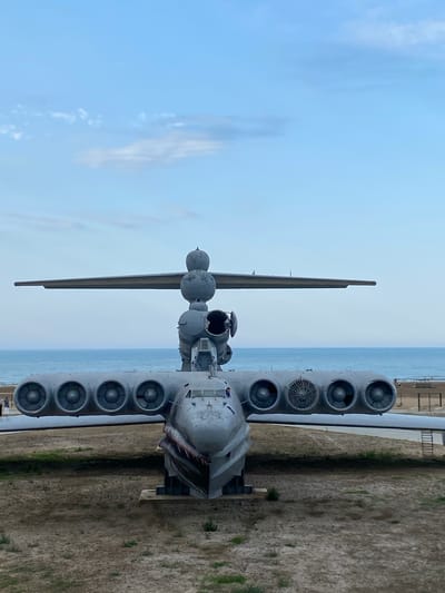 A front-facing, eye-level, full shot captures an imposing, gray, Soviet-era ekranoplan (ground-effect vehicle) resting on a sandy, dry dirt surface. The colossal aircraft features a distinctive design with a long, wide body and two expansive wings that stretch horizontally. Along the underside of the starboard wing, six circular engine nacelles are visible, with a seventh engine, a different design, positioned further outboard. The port wing mirrors this arrangement, though fewer engines are fully visible due to the angle. The nose of the ekranoplan is pointed and features a painted shark mouth graphic with sharp teeth. A tall, multi-tiered structure rises from the center of the aircraft's body, housing additional engines and what appears to be a cockpit area. The background is dominated by a vast, calm ocean stretching to the horizon, with a hazy, pale blue sky above, marked by thin, wispy clouds. The environment suggests a coastal location, possibly a museum or preserved exhibit.