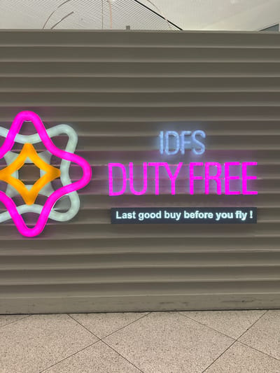 This media file captures an indoor commercial scene, likely a duty-free shop within an airport, given the prominent signage and the provided location context of Nouaceur, Morocco (which hosts Casablanca's Mohammed V International Airport).

The main subjects are several brightly lit neon signs set against a light gray, vertically ribbed wall. On the left, the letters "IDFS" are illuminated in blue-white neon, oriented vertically downwards. To its right, the words "DUTY FREE" glow in vibrant pink neon, also oriented vertically downwards. Further to the right, a black rectangular sign with white text reads "Last good buy before you fly !", with the text oriented vertically upwards.

Below the text-based neon signs, a decorative neon element is present. This abstract design features intertwined, multi-layered shapes resembling a star or flower, rendered in pink, white-blue, and yellow-orange neon tubes.

Portions of a light-colored, speckled ceiling are visible in the top left of the image, while a light-colored, speckled tiled floor with dark grout lines can be seen in the bottom right. The overall lighting is artificial, provided by the neon signs, creating a modern and inviting commercial atmosphere. No people or specific activities are depicted; the image focuses solely on the architecture and signage of the retail space.