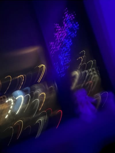 Blurred, colorful lights adorn a dark background. Patches of blue and purple lights cluster in the upper left corner. Arcs of yellow, white, and red streaks suggest motion. The overall effect is abstract and impressionistic, hinting at a dynamic scene with light and movement, possibly inside a room or at an event.