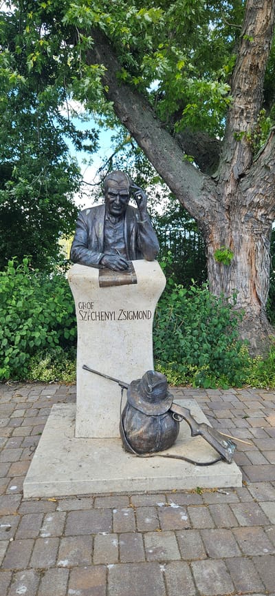 The image shows a bronze statue of a man in a suit, seated and holding a pen to a notepad, with his hand on his head. The statue is mounted on a stone pedestal with the inscription "Gróf Széchenyi Zsigmond" in Hungarian. In front of the pedestal, there is a bronze sculpture of a rifle and a hat resting on a bag. The scene is set outdoors, with lush green foliage and a large tree in the background. The ground is paved with interlocking bricks. This memorial is located in the town of Balatongyörök, Hungary, on the northern shore of Lake Balaton.