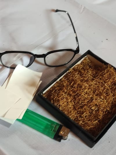 The scene, located in Staufenberg, Germany, depicts several objects arranged on a light-colored, textured surface, possibly fabric. In the upper left quadrant, a pair of black-framed spectacles rests partially unfolded, with the right temple arm extending upwards. To the right, slightly below the center, is an open, rectangular black container filled with finely shredded, light brown organic material, consistent with loose-leaf tobacco. Below the spectacles and to the left of the tobacco container, a stack of off-white, rectangular paper sheets, consistent with rolling papers, is fanned out. A translucent green disposable lighter with a metallic top mechanism is positioned horizontally below the rolling papers and to the left of the tobacco container. No individuals, actions, or specific environmental identifiers beyond the immediate surface are visible.