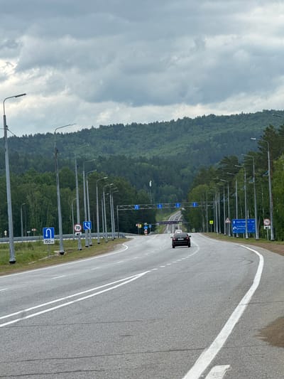 The image shows a cloudy day on a multi-lane highway leading into a forested mountain range. The highway has distinct white lane markings and is bordered by a metal guardrail on the left and a line of streetlights on the right. Several road signs are visible, including a speed limit sign of 70, a U-turn sign with a distance of 150 meters, and directional signs indicating "Листвянка" (Listvyanka) and "Иркутск" (Irkutsk). A dark sedan is driving away from the camera on the right lane. The landscape is dominated by dense green forest that covers the hills. The presence of highway infrastructure and signs with Russian place names suggests this location is on a major road in or near Patrony, Russia.