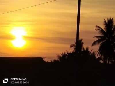 The image shows a sunrise or sunset with a bright orange-yellow sky. The sun is partially obscured by clouds or haze. In the foreground are silhouetted shapes of trees, including palm trees, and what appears to be a building roof. A utility pole is present on the right side of the frame. The location is Cot Buket, Indonesia, indicated by the camera settings in the lower-left corner of the image. The time is 06:31 on 2026.05.23 according to the image's timestamp.