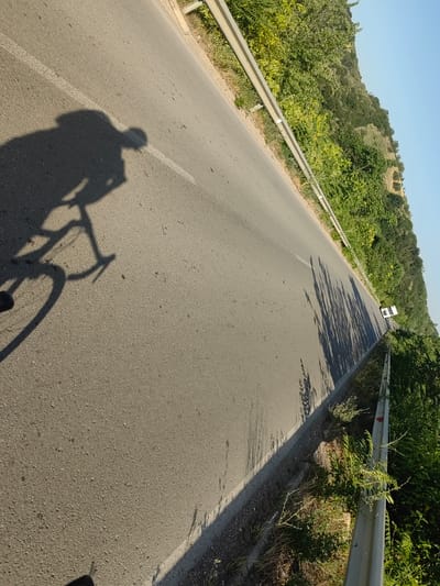 The image captures a paved road in Staliyska mahala, Bulgaria, with a guardrail on the right and lush green vegetation lining the roadside and extending up a gentle slope. The perspective is from a moving vehicle, likely a bicycle, evidenced by the prominent shadow of a cyclist and their bike cast on the asphalt. The cyclist's shadow, detailed and sharp, shows a person in riding posture with distinct handlebars and wheels. In the distance, a white van is visible driving on the road. The sky is a clear, pale blue, indicating a sunny day. The overall scene depicts a tranquil journey through a scenic, rural-like area within the city.