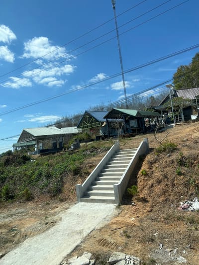 A sideways view shows a hillside with structures featuring green roofs. A set of concrete stairs with handrails ascends the slope, adjacent to a paved walkway. Power lines are visible against a blue sky with scattered clouds. The surrounding terrain consists of dry grass and sparse vegetation. The composition is framed with an emphasis on the architectural elements and the surrounding natural environment.