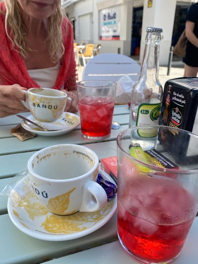 A close-up shot captures two cups of coffee and two red drinks with ice on a light green table, suggesting an outdoor cafe setting in Conil de la Frontera, Spain. In the foreground, a white coffee cup with yellow leaf-like designs and the text "BANDU" sits on a matching saucer. A small spoon rests on the saucer, and a sugar packet is partially visible. To the right, a red drink in a frosted glass is filled with ice. Behind these, another white cup, presumably also with coffee remnants, is partially visible, and a second red drink in a glass is to its right.
A clear glass bottle with a metallic cap, labeled with "33 cl" and "Cerveza," stands between the cups. To its right is a black napkin dispenser with the "Estrella Galicia" logo, indicating a possible connection to a brewery or bar. In the background, a woman wearing a red shawl over a white top is visible, her hands holding one of the coffee cups. The setting appears to be a street-side cafe, with blurred white buildings and tables and chairs in the distance, consistent with the architecture and atmosphere of Conil de la Frontera. The lighting suggests daytime.