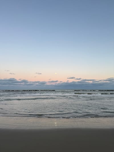 This image captures a twilight coastal scene in Grottammare, Italy. The sky exhibits a gradient, transitioning from a light blue at the zenith to a soft pink and orange along the horizon. A horizontal band of scattered, flat gray clouds stretches across the mid-sky. A prominent, pale yellow-white moon is visible in the upper mid-right quadrant. The sea in the midground features muted grey-blue water with gentle, low-amplitude waves. Multiple dark, linear rock breakwaters are positioned horizontally across the water, extending from left to right. The foreground consists of a damp, dark sandy beach, displaying a faint vertical reflection of light near the waterline, consistent with the moon's illumination. No individuals or man-made objects, beyond the breakwaters, are present within the frame.