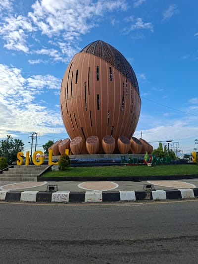This is a medium-angle, eye-level, outdoor, full shot photograph of the Sigli roundabout in Lampeudeu Baroh, Indonesia, under a partially cloudy blue sky. The focal point is a large, bulbous structure with a dark, patterned dome, resembling a seed or fruit, constructed with vertical copper-colored panels. The lower portion of the structure is adorned with several smaller, similar bulbous shapes. In front of the main structure, large yellow block letters spell out "SIGLI." To the right of the main structure, a green and red logo with the text "Bank Aceh" is visible. The roundabout is surrounded by a neatly manicured green lawn, with a paved road and a curb in the foreground. Electrical poles and trees are visible in the background.