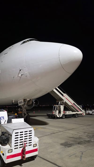 The image shows the nose of a large white airplane on an airport tarmac at night. A mobile staircase, attached to a truck, is positioned against the side of the aircraft, suggesting boarding or disembarking activities. The tarmac is concrete with yellow markings and various airport equipment is visible, including a tow tractor positioned in the foreground. The lighting suggests it's nighttime, possibly with overcast weather, as the sky is dark and there are no stars visible. The overall scene is typical of an airport at night, with the focus on the aircraft and the equipment servicing it. Visible text includes "BAQ" on a placard near the aircraft's door, and "PS 12" on the side of the staircase truck. The setting is an airport tarmac in Fujairah, United Arab Emirates.