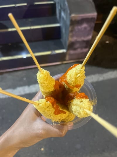 A close-up, eye-level, over-the-shoulder shot shows a person's hand holding a clear plastic cup filled with four deep-fried snacks on wooden skewers, drizzled with a red sauce. The snacks are golden yellow and appear to be oblong or slightly irregular in shape, with a textured, crispy exterior. The red sauce is thick and appears to be a chili or ketchup-based sauce. The background is blurred, showing a dark asphalt surface, possibly a street or pathway, and a set of dark steps with purple and yellow stripes. The lighting suggests it is evening or nighttime. The scene is indicative of a street food vendor in Tamanarum, Indonesia, given the style of food and typical urban street setting.
