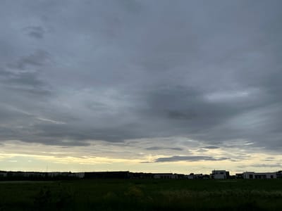 The image captures a wide, horizontally oriented view of a suburban or rural landscape under a dramatic, cloudy sky. In the foreground, a dark green field with sparse, low-lying vegetation and some wildflowers stretches across the bottom of the frame. In the middle ground, a line of buildings forms the horizon. These buildings appear to be a mix of dark, elongated structures and lighter, modern residential houses with flat roofs, situated along a road. The sky dominates the upper two-thirds of the image, filled with a dense, overcast layer of gray clouds. Towards the horizon, a sliver of pale yellow and white light suggests the presence of the sun or a break in the clouds, casting a soft glow on the lower sky and the upper portions of the buildings. The overall atmosphere is somber and tranquil, typical of an overcast day, possibly near dusk or dawn. There are no people or animals visible in the scene. The location is identified as Harkujärve, Estonia.