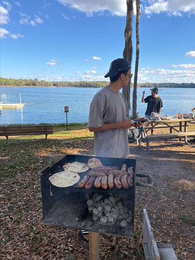 The image depicts an outdoor scene at a lakeside park in Micanopy, United States, under a clear blue sky with scattered white clouds. In the foreground, a black, square charcoal grill contains approximately 18-20 browned sausages arranged in rows, two flatbreads, and one bread roll, all cooking over grey charcoal briquettes.

A young adult male stands in profile to the right of the grill, wearing a light grey oversized t-shirt, a dark baseball cap with a white emblem, and sunglasses tucked into his collar. He holds a small, dark rectangular object in both hands. Behind him and to the right, another young adult male stands behind a wooden picnic table. This second individual wears a dark t-shirt and a dark baseball cap with sunglasses on his face, and his right arm is raised with the index finger pointing upwards. Various items, including a bottle with a red cap, are visible on the picnic table.

The background features a wide, calm blue lake with a tree-lined far bank. A white floating dock with a ladder is present on the left side of the lake. A red and white "No Diving" sign is posted on a pole near the water's edge. Numerous small boats are visible on the lake in the distance. The ground in the foreground is covered with dry leaves and exposed dirt. A tall, slender tree with Spanish moss hanging from its branches stands in the mid-ground. A wooden park bench is positioned near the water on the left.