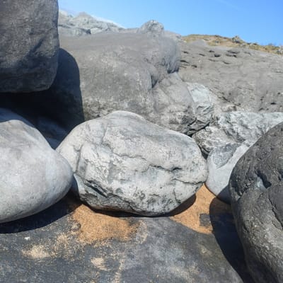 This is a close-up, ground-level shot of large, weathered rocks on a sandy beach. The rocks are varying shades of gray and appear smooth and rounded, suggesting they have been shaped by water and time. Some rocks are partially covered by fine, light-brown sand. The lighting suggests a sunny day, with strong shadows cast by the rocks. In the background, the terrain rises with more rocks and sparse vegetation, leading towards a bright blue sky. This scene is consistent with the rocky coastline found in Fanore, Ireland. There are no people or discernible actions in the image.