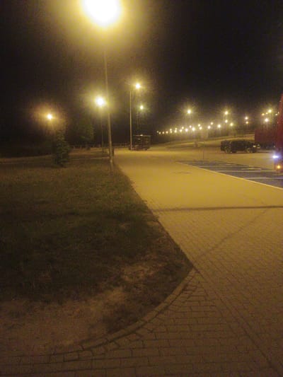 The image depicts a nighttime scene at a rest stop or parking area in Bratkowice, Poland. A bright street lamp illuminates the foreground, casting a yellow glow on a paved walkway made of interlocking bricks. To the left of the walkway, a grassy area is visible, with a small tree in the middle ground. In the background, more street lights line a paved road. Several vehicles are parked in the distance, including a dark-colored SUV and the rear of a red truck. The overall impression is one of a typical, well-lit roadside facility at night.