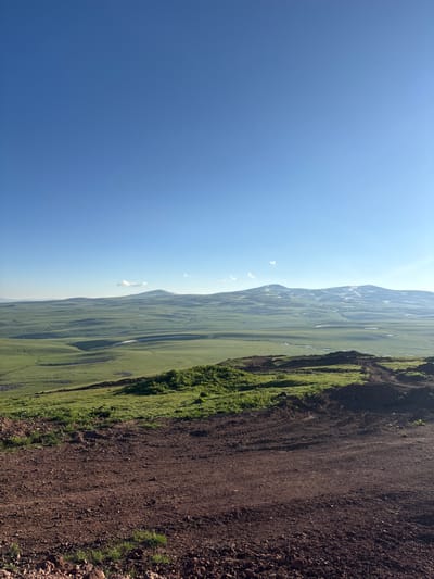 The image captures a panoramic view of a vast, undulating landscape under a clear blue sky. In the foreground, a reddish-brown dirt path, possibly a road or a hiking trail, leads the eye into the scene. The path is uneven and textured with small stones and tire tracks, suggesting recent activity.
Beyond the dirt path, lush green rolling hills dominate the midground. These hills are contoured by gentle slopes and shadows, creating a sense of depth and vastness. A winding river or stream can be faintly seen snaking through the valleys of these green hills.
In the distance, a range of mountains rises, some peaks still bearing patches of snow. The mountains appear hazy, possibly due to atmospheric conditions or distance. A few small, wispy clouds are scattered across the sky, particularly near the mountain range.
The overall impression is one of serene, natural beauty, characteristic of the highlands around Jermuk, Armenia. The scene appears to be an outdoor vista, possibly a viewpoint overlooking the mountainous terrain for which Jermuk is known. No people or specific objects of interest, beyond the natural landscape, are visible. The lighting suggests it is either early morning or late afternoon, with the sun casting long shadows.