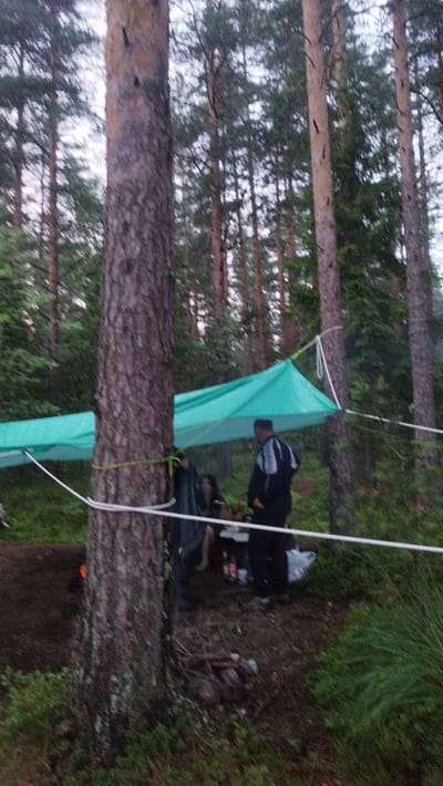 The image depicts a forest scene with a group of people gathered under a turquoise tarp, which is suspended between trees. The tarp appears to be set up as a shelter. In the foreground, a large tree trunk dominates the left side of the frame, with its bark textured and weathered. A green rope is tied around this tree. Several other trees, characteristic of a pine forest, are visible in the background, with their trunks and needles creating a dense canopy.
Three individuals are visible beneath the tarp. One person, wearing a dark jacket with white accents, stands to the right of the group, facing away from the camera. Another person, partially obscured by the tarp, is seated to the left. A third individual, also seated and mostly hidden, is near the center of the group. A table or low surface is set up in front of the seated individuals, with various items on it, including bottles and bags. The ground beneath the tarp is covered with forest floor debris, including dirt and vegetation. A white rope is strung horizontally across the middle of the frame, in front of the tarp.
The overall impression is of an outdoor gathering or campsite in a wooded area. The lighting suggests it may be overcast or dusk. There is no indication of the location being in the city of Tarasovskoe, Russia, as the provided context states.