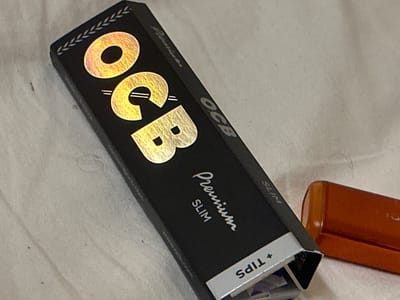 A close-up, high-angle shot shows a black OCB Premium Slim rolling paper booklet. The booklet is partially open, revealing the silver foil lining and some contents inside. The OCB logo, in a metallic gold font, is prominently displayed on the lid of the booklet. Below the logo, "Premium" is written in a stylized script, followed by "SLIM" in a block font. The booklet is resting on a textured, light-colored surface, possibly a tablecloth or a neutral fabric. To the right of the booklet, a small, orange, cylindrical object, possibly a lighter or a container, is visible. The image is well-lit, with the metallic elements on the booklet reflecting the light. There is no discernible urban environment or location within Hradec Králové, Czechia.
