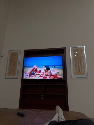 The image depicts a television screen displaying two women lounging on a beach. The woman on the left has pink hair and is wearing a blue bikini top. The woman on the right has dark hair and is wearing a bikini top. They are both lying on red and white striped towels. A pink radio is visible next to the woman on the left. The television is mounted in a dark wood entertainment center. On either side of the entertainment center are framed abstract art pieces. In the foreground, a person's legs wearing white socks are visible, resting on a brown surface, likely a bed or couch. A black remote control is on the surface near the person's feet. The scene appears to be indoors, with a neutral-colored wall in the background. The location of Santiago Etla, Mexico is not evident in the image.