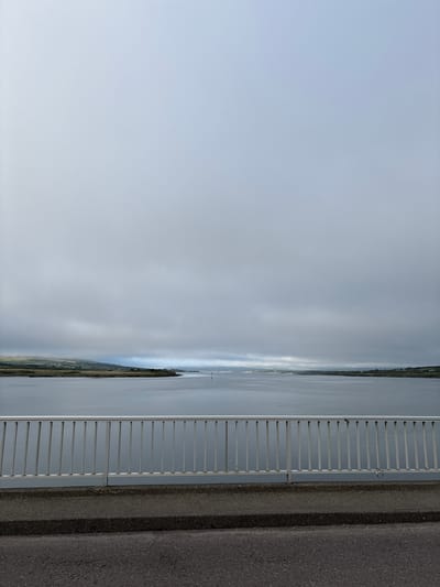 This image captures a tranquil scene of a wide body of water, likely a bay or estuary, under a heavily overcast sky. In the foreground, a silver metal railing runs horizontally across the frame, suggesting the viewpoint is from a bridge or elevated walkway. Below the railing is a concrete barrier, and further down, a dark asphalt surface, possibly a road. The water is calm and reflects the muted light of the sky. In the distance, a low-lying, green landscape with some scattered structures can be seen along the far shore. The overall atmosphere is one of subdued light and quietude, characteristic of a cloudy day in a coastal region. The location is identified as Portmagee, Ireland.