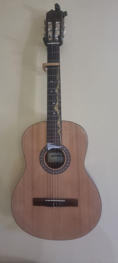 This image displays a classical acoustic guitar mounted horizontally on a textured, light-colored wall. The guitar is positioned so that the headstock is on the left and the body is on the right. The guitar has a light wood finish with a dark wood bridge and fretboard. The soundhole is circular and decorated with an intricate rosette pattern. A label with the brand name "admira" is visible inside the soundhole, along with smaller text that appears to be a model name. The fretboard has decorative inlay markings that resemble mountain peaks. A wooden capo is attached to the neck of the guitar just above the nut. The tuning pegs on the headstock are white. The overall setting appears to be indoors, likely a home or practice space, given the wall texture. There are no people or any immediate activity suggested in the image, making it a static display of the instrument. The lighting is even, suggesting it is daytime or well-lit indoors, with no discernible weather conditions. The location context provided is Chernomorskiy, Russia.