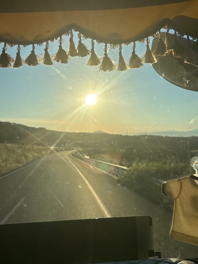 The image shows a view from inside a vehicle, looking out through the windshield. The sun is setting, casting a warm glow over a winding road that leads into a hilly landscape. The sky is a clear blue, with the sun positioned in the upper center of the frame. A decorative fringe with tassels hangs from the top of the windshield. In the foreground, a dark dashboard is visible, and a light-colored t-shirt is hanging from a hook on the right side of the windshield. The location is identified as Avra, Greece.