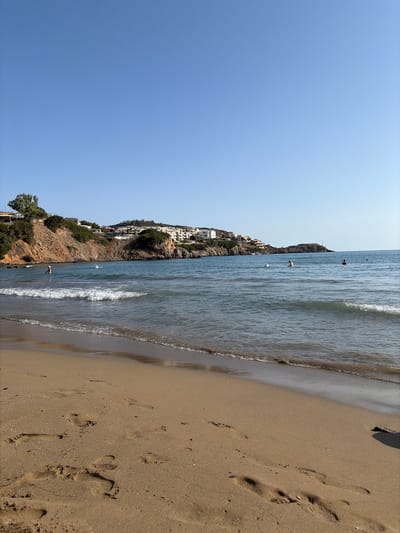 The image depicts a sunny day at a beach in Vlichada, Greece. The foreground is dominated by a sandy beach with visible footprints, indicating recent human activity. Gentle waves lap the shore, creating a white foam line along the wet sand. In the middle ground, the calm, blue sea stretches towards the horizon. Several individuals are visible in the water, some swimming and others standing. In the background, a rocky coastline rises, dotted with green vegetation and a cluster of white buildings, characteristic of coastal architecture in Greece. The clear blue sky above completes the serene beach scene.