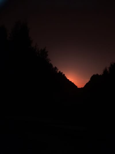 A silhouette of trees frames a dark sky at dusk. The horizon is illuminated by a faint orange glow, suggesting the presence of the sun below the visible horizon. The scene is characterized by deep shadows and a muted color palette, with the darkness of the trees contrasting with the subtle light of the sky. There are no people, objects, or visible actions depicted in the image. The environment appears to be natural, with the trees suggesting an outdoor setting, possibly a rural or wooded area. The image is devoid of any specific landmarks that would definitively place it within the city of Sergach, Russia.