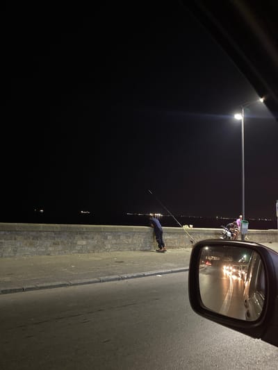 The image captures a nocturnal scene near the Beau Fraisier, Algeria. A person is seen fishing with a rod, leaning against a stone wall along a waterfront. Further down the wall, two individuals are seated on a scooter, and another person stands nearby, illuminated by a street lamp. The dark expanse of the sea stretches out, dotted with distant lights from ships and possibly the shoreline. The foreground shows a road, partially reflected in a car's side mirror, revealing oncoming headlights and the glint of wet asphalt. The overall atmosphere is one of quiet evening activity by the water.