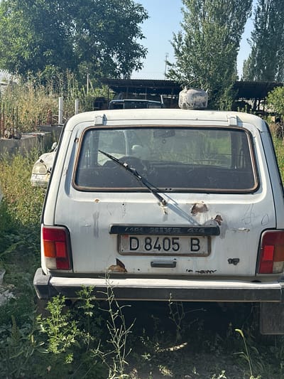 A white, older model Lada Niva 4x4 is parked outdoors in an overgrown lot, likely in International, Kyrgyzstan. The car is rear-facing, with a license plate reading "D 8405 B". It appears weathered and rusty, with some of the paint peeling off and a notable amount of rust on the rear of the vehicle. A windshield wiper is visible on the rear window. In the background, there are trees, some fencing, and other parked vehicles, suggesting a junkyard or an area where old cars are stored. The overall environment is natural, with tall weeds and grass growing around the car. A metal teapot sits on the roof of the car.