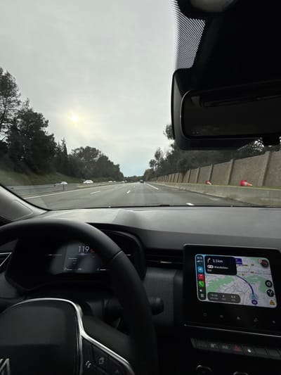 The image depicts a first-person view from the driver's seat of a vehicle traveling on a multi-lane highway, specifically the A8 autoroute in the vicinity of Mougins, France.

The exterior scene shows a moderately trafficked highway under an overcast sky with a bright sunburst visible through the clouds. On the left, an embankment is densely covered with dark green trees. On the right, a tall, segmented beige sound barrier or retaining wall runs alongside the highway, with additional trees visible above it. Two red and white traffic cones are partially visible on the right shoulder, leaning against the base of the wall.

Inside the vehicle, a modern dashboard with a dark, textured finish is visible. The digital instrument cluster, partially obscured by a black multi-function steering wheel with chrome accents, displays a speed of "119 km/h" and "250 km" (likely range). A central infotainment screen shows a navigation map indicating a destination of "Mandelieu-Est" 3.3 km away, with an estimated arrival time of "15:24" and the current time as "15:15." A "5G" signal icon is also present on the screen. The vehicle's interior rearview mirror is mounted to the windshield, which features a dotted sensor area behind it. No occupants are explicitly visible, but the driver's perspective implies operation of the moving vehicle.