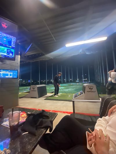 The image depicts an indoor Topgolf venue at night. A person dressed in black is swinging a golf club on the green. A Topgolf kiosk and monitor are visible nearby. Other patrons are seated in the foreground, one in a white sweatshirt. There's a black marble table with napkins and cups. In the background, a bright fluorescent light illuminates the metal roof. Two screens with bright graphics are displayed at the lower left corner. Golf targets and netting stretch far into the background.