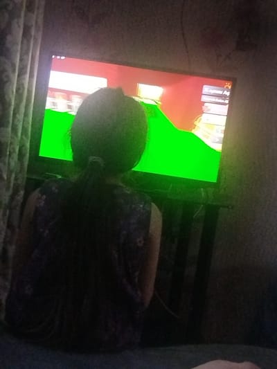 A person is viewed from behind, facing a television screen, in a dimly lit room. The television displays a colorful video game with large areas of bright green. The person has dark, curly hair pulled back in a ponytail and is wearing a dark, patterned top. The room appears to be a living space, with a curtain visible on the left side of the image and a dark wall behind the television. The overall scene suggests a domestic setting where the person is engaged in playing a video game. The image does not contain any elements that would allow for the determination of its location in the city of Sabunchu, Azerbaijan.