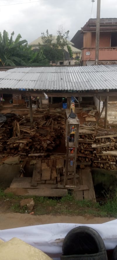 The image depicts an outdoor scene in Ezinifite, Nigeria, under an overcast sky. In the midground, a rudimentary structure with a corrugated metal roof supported by wooden posts covers an area used for storing and selling materials. Beneath the roof, extensive piles of cut firewood and logs are visible, with some stacks neatly arranged and others forming a heap. A wooden stand displays multiple plastic jerrycans, predominantly yellow and red, likely containing fuel, alongside some blue containers. The ground beneath the structure is exposed earth.
In the background, several residential buildings with various roof types are present, partially obscured by lush green vegetation, including what appears to be banana trees. One two-story building on the right features an orange-colored facade with an upper balcony and a white rectangular sign bearing the number "136." A utility pole with an attached light fixture stands on the right, extending above the buildings. The foreground shows brown soil with patches of green grass, a small rock, and a white tarpaulin or fabric partially covering an object, with a black tire visible on the far right. No individuals are discernible or actively engaged in specific tasks.