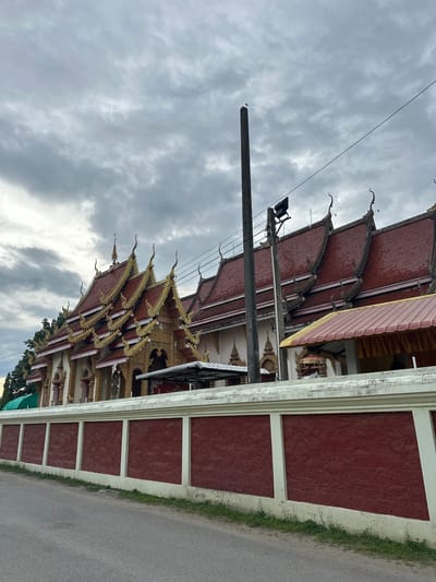 The image shows a temple in Ban Patan, Thailand, under a cloudy sky. The temple features a red tiled roof with ornate golden trim, and a tall, red brick wall with white accents separates it from the street. A concrete utility pole stands in front of the temple, with a bird perched on top. The sky is overcast with grey clouds. The scene is peaceful and somewhat subdued due to the weather.