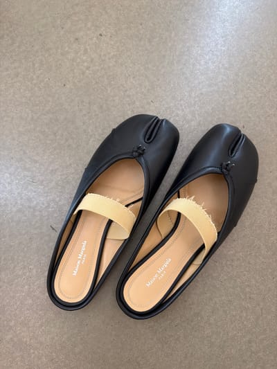 This media file features a pair of black Maison Margiela tabi mules, presented from a top-down perspective. The shoes are placed on a textured, light-colored surface that appears to be concrete or a similar material. One shoe is positioned slightly above and to the right of the other. Both shoes are characterized by their distinctive split-toe design and a tan elastic strap across the instep. The interior of the shoes reveals the "Maison Margiela PARIS" branding printed on the insoles. The lighting suggests an indoor setting, possibly a room with overhead illumination, casting subtle shadows. There are no people visible in the image, and the focus is solely on the footwear. The scene is static, with no activity taking place. The overall impression is clean and minimalist, highlighting the design of the shoes.