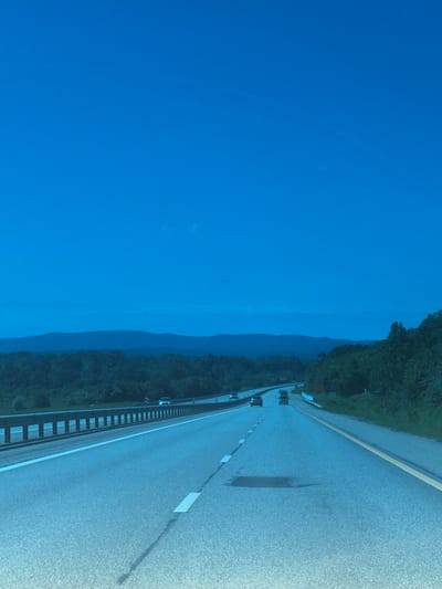 The image depicts a view from a moving vehicle on a highway. The scene includes a multi-lane road with painted white lines marking lanes, a metal guardrail separating the road from the adjacent foliage, a clear blue sky, and a distant mountain range. There are other vehicles visible on the road ahead. Based on the information provided, the location is Saratovskaya, Russia. The image conveys a sense of movement and travel through a rural or scenic area.