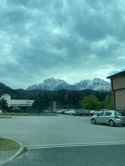 The scene depicts an outdoor environment in Jablanica, Bosnia and Herzegovina. The background is dominated by a range of snow-capped mountains under a light blue-grey, heavily overcast sky.
In the midground, a paved asphalt parking lot extends across the frame. It contains multiple parked passenger vehicles, primarily light-colored sedans and hatchbacks (silver, white, grey). A yellow taxi is identifiable among the vehicles on the right side of the lot. Two blue circular traffic signs, including one indicating a mandatory direction, are visible adjacent to the parked cars.
To the left of the parking lot, a long, light-colored building with a dark roofline is present, featuring a prominent cylindrical tower-like structure at its left end. Several evergreen trees and one deciduous tree with visible green foliage are situated between this building and the parking area. A multi-story building with a light brown facade and a dark roof is partially visible on the far right edge of the frame, adjacent to the parking.
The immediate foreground consists of a grey paved road surface. A curved green grass verge and a concrete curb are visible on the far left of the foreground. No individuals are present within the depicted scene. The illumination is consistent with diffused daylight under an overcast sky.