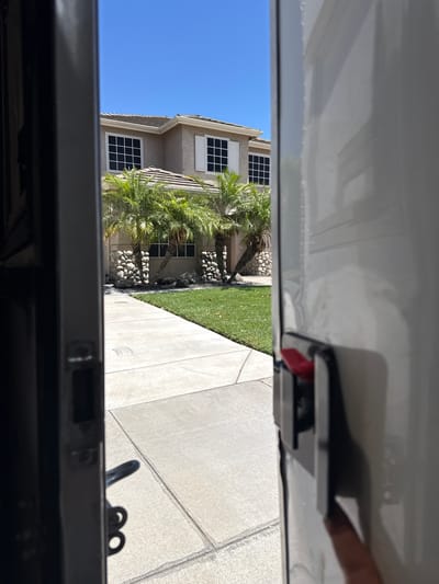 The image, taken on a sunny day in Ripon, California, shows a view from inside an open doorway, likely of a vehicle or storage unit. The perspective looks outward towards a two-story house with a light-colored stucco exterior, white-trimmed windows, and a shingled roof. The house is surrounded by a neatly manicured green lawn and several palm trees. In the foreground, a concrete sidewalk leads away from the viewer towards the house. A black door frame is visible on the left, and the white side of what appears to be a vehicle or storage unit dominates the right side of the frame. A red latch mechanism is visible on the white surface. The sky is clear blue.